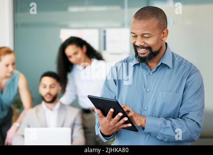 Were in for a busy day. a mature businessman standing in the office and using a digital tablet while his colleagues work behind him. Stock Photo