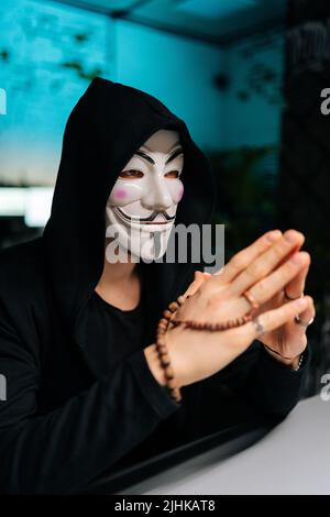 Close-up vertical shot of dangerous hacker man wearing anonymous mask and hoodie holding beads in hands and looking away sitting at table in basement Stock Photo