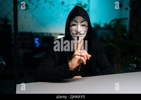 Portrait of unrecognizable hacker man wearing anonymous mask and hoodie warningly gesturing with hands sitting at table in basement room. Stock Photo