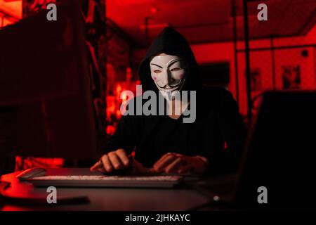Portrait of dangerous unrecognizable hacker man wearing anonymous mask starting breaking computer system and typing password for hacking bank account. Stock Photo