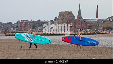 Portobello Beach, Edinburgh, Scotland, UK. 19th July 2022. Cloudy hot Sultry afternoon at the seaside with temperature around 27 degrees centigrade for the crowds who visited the east coast beach. Pictured: A young couple heading to the shore of the Firth of Forth with their paddleboards.  Credit: Arch White/alamy live news. Stock Photo