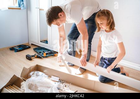 Dad,little girls daughters assemblle wooden baby bed according to instructions in kids room. Helping father, mounting children's furniture. Self-assem Stock Photo