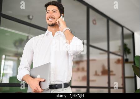 Ambitious handsome hispanic male entrepreneur, start-up owner in white shirt standing with laptop in modern office and talking on phone, man has phone conversation with partner, solving business tasks Stock Photo