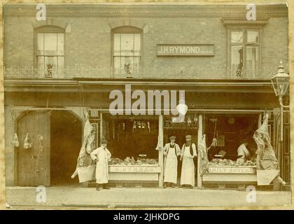 Original Edwardian era photograph of  J.H. Rymond family butchers, group of butchers assistants / young apprentices,  selling sides of beef displayed outside and pickled tongues advert on the window., Harvest Festival poster in the window, probably located in Warwickshire, England, U.K. circa 1908. Stock Photo