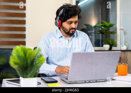 Young man with wireless headphones working on laptop while sitting at office - concept of online meeting, working lifestyles and employment Stock Photo