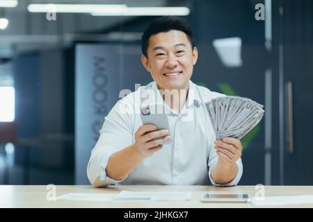 Successful asian businessman working in office, man smiling and looking at camera, holding phone and money dollars cash Stock Photo