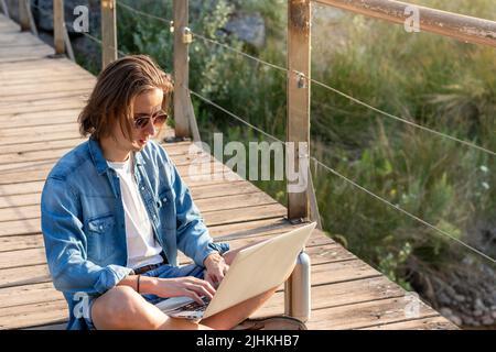 Young online working as freelancer man with long hair sitting on the floor woking on his laptop while on vacation Stock Photo