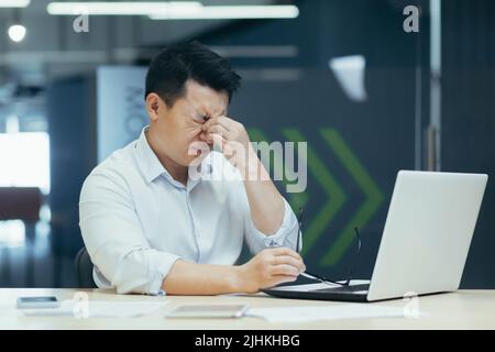 Tired Asian businessman working late in office, sore eyes, man with laptop Stock Photo