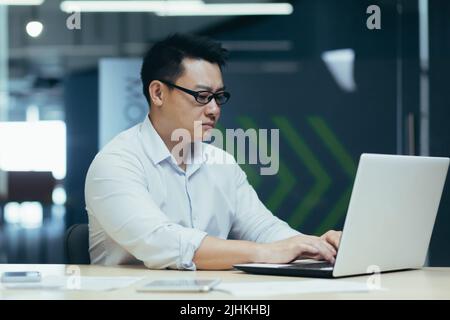 Serious asian boss in glasses working and typing on laptop, man concentrated in modern office Stock Photo