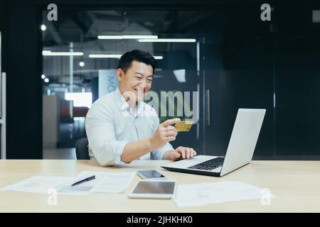 Happy Asian businessman working in modern office, man making online money transfer, using laptop and holding bank credit card Stock Photo