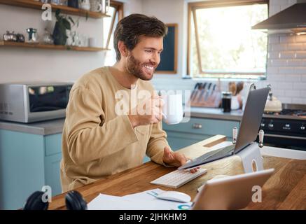 Happy young caucasian man drinking coffee while working on laptop in kitchen, checking his email or searching information while doing freelance work Stock Photo