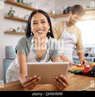 Portrait of beautiful young woman browsing on digital tablet while her boyfriend cooks in the background. Young hispanic female searching for recipe Stock Photo