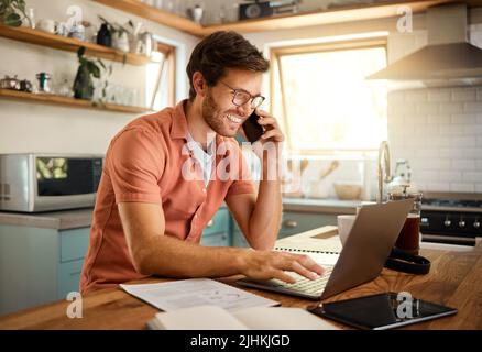 Young content caucasian businessman wearing glasses on a call using a phone while working on a laptop at home alone. Happy male businessperson smiling Stock Photo