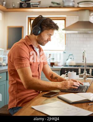 Young serious caucasian businessman wearing headphones and listening to music while using a laptop at home alone. One male businessperson typing on a Stock Photo