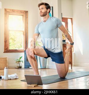 One fit young caucasian man stretching legs with lunge bodyweight exercise and holding foot while training with online tutorial on digital tablet at Stock Photo