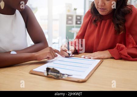 Two unknown ethnic business woman sitting together and signing office contract. African american professional using hand gesture to point on paperwork Stock Photo
