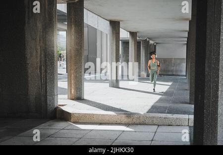 Young hispanic female athlete wearing headphones while running though a building outside. Young sportswoman focused on her speed, body, fitness and Stock Photo