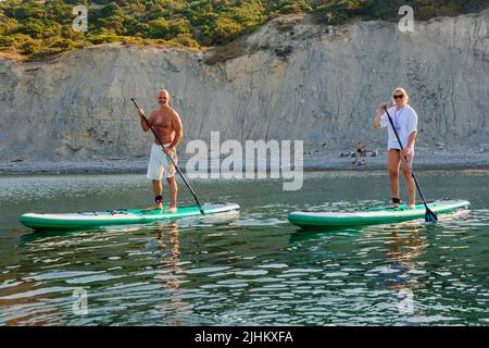 July 5, 2022. Dalaman, Turkey. Older couple on paddle board on quiet sea with rocky coastline and morning sunlight. Active lifestyle for aged man and Stock Photo