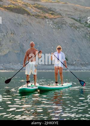 July 5, 2022. Dalaman, Turkey. Older couple on paddle board on quiet sea with rocky coastline and morning sunlight. Stock Photo