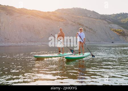 July 5, 2022. Dalaman, Turkey. Older couple on paddle board on quiet sea with rocky coastline and morning sunlight. Active lifestyle for aged man and Stock Photo