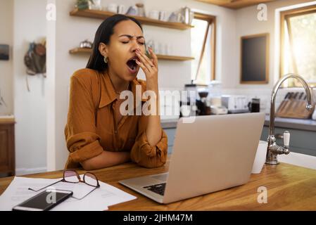 Bored mixed race businesswoman yawning while going through paper and bills and working on a laptop at home. Tired hispanic female businessperson Stock Photo