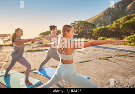 Diverse smiling yoga women in warrior pose during outdoor practice in remote nature. Group of happy active friends using mat and balancing while Stock Photo