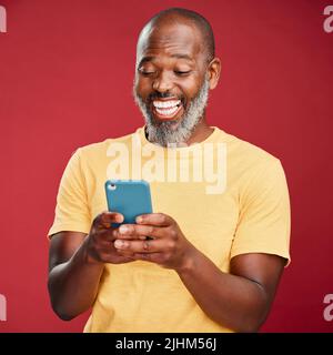 One happy African American man standing against a red studio background, holding and using his cellphone to browse the internet. Smiling black man Stock Photo