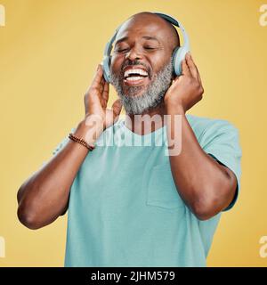 Happy mature African American man standing alone against a yellow background in a studio and wearing headphones to listen to music. Smiling portrait Stock Photo