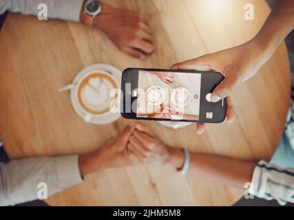 Close up of device screen while couple holds hands with two cups of coffee on a wooden table while taking a selfie on smartphone. Female and male Stock Photo
