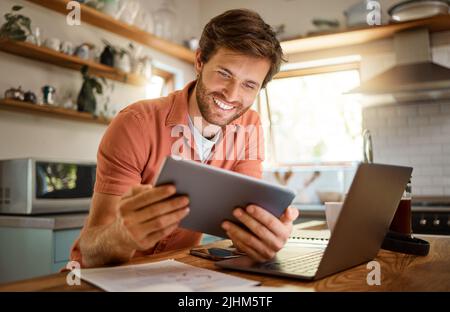 Young cheerful caucasian businessman using a digital tablet while working on a laptop at home alone. Happy male businessperson smiling and using Stock Photo