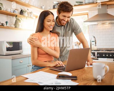 Young happy interracial couple bonding while working on a laptop together at home. Caucasian boyfriend and girlfriend planning and using a laptop Stock Photo