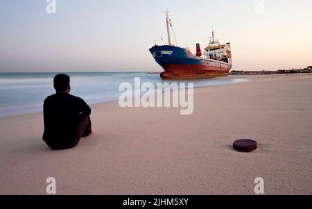 Middle aged man sitting on the beach and watching the shored ship in Sharjah, United Arab Emirates Stock Photo