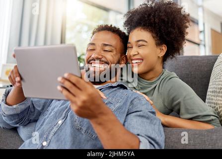 Mixed race couple smiling while using a digital tablet together at home. Joyful hispanic boyfriend and girlfriend laughing while relaxing and using Stock Photo