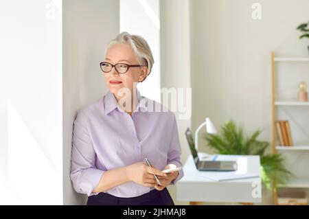 Smiling senior business lady with mobile phone in hands looking into distance through window. Stock Photo