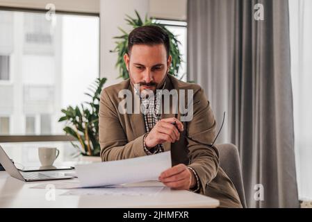 Serious pensive thoughtful focused young business accountant bookkeeper in office looking at papers and documents and working with laptop and income t Stock Photo