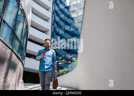 Low angle view of the positive entrepreneur walking with smart phone. Young business professional is carrying business bag. He is going on footpath ag Stock Photo