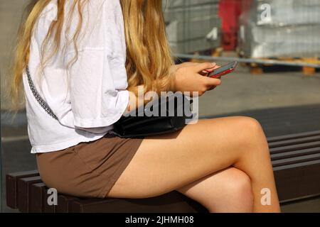 Girl with long fair hair wearing shorts sitting on wooden bench with smartphone in hands, summer leisure in city Stock Photo