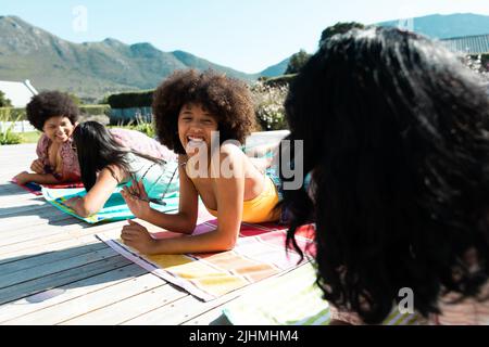 Cheerful biracial female friends talking while relaxing on towels at poolside during summer Stock Photo
