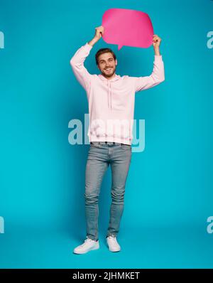 Image of emotional excited young man posing isolated over light blue wall background holding speech bubble Stock Photo