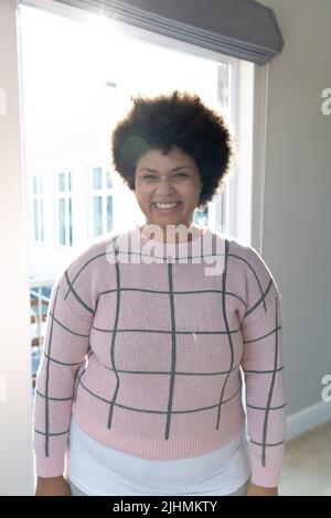 Smiling biracial young woman with afro hair standing against sunlight streaming through window Stock Photo