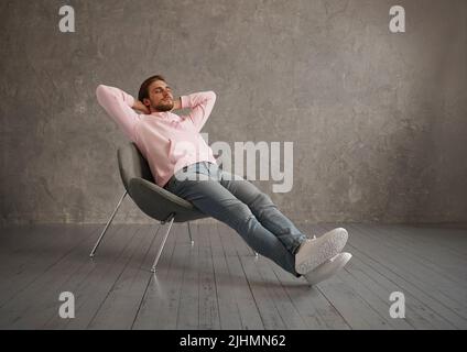 Millennial Guy Relaxing Sitting In Comfortable Chair Holding Hands Behind Head Resting With Eyes Closed Over Gray Wall Stock Photo