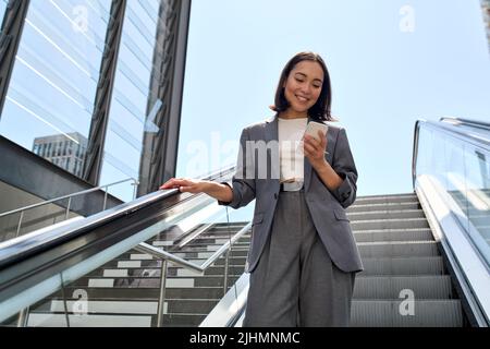 Smiling young Asian business woman standing on escalator using phone. Stock Photo