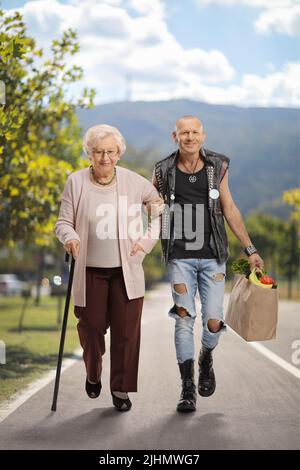 Man helping a senior woman with a walking cane and carrying a grocery bag outdoors Stock Photo