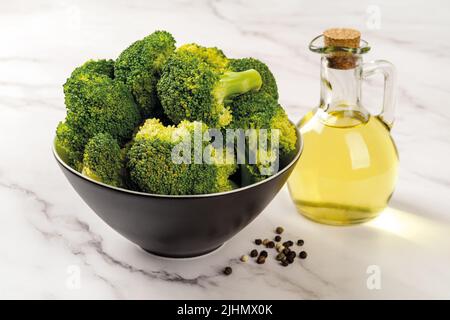 Fresh broccoli and olive oil on the marble countertop. Raw green broccoli florets in a black bowl and glass jug of sunflower oil. Low calories food. Stock Photo
