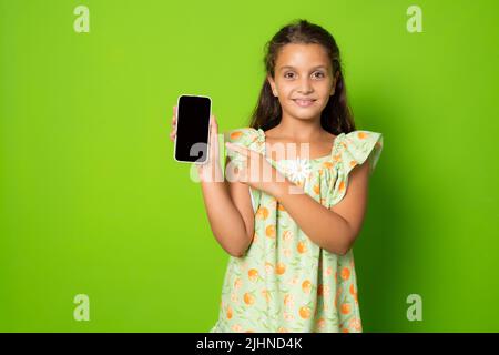 Little girl showing a smart phone screen standing isolated over green background. Stock Photo