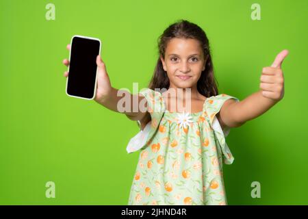 Little girl showing a smart phone screen and thumb upstanding isolated over green background. Stock Photo