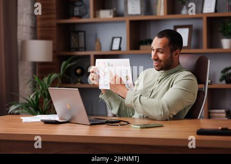 Young smiling mixed race businessman have video call sitting in office, showing financial report with graphs and charts to colleagues, partners and Stock Photo
