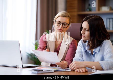 Two businesswomen discussing financial report, holding documents with graphs and charts, communicate before meeting with investors at office Stock Photo