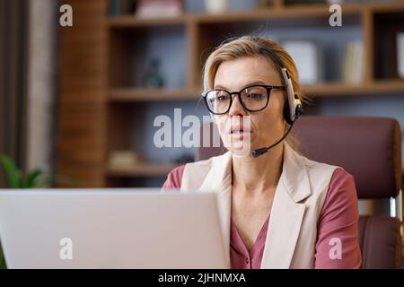 Smiling businesswoman wearing headset talking to laptop web camera sitting in office. Recording vlog, online video conference, video call, online Stock Photo