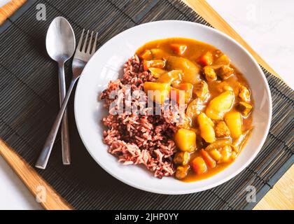 Japanese curry with rice berry in round white plate, healthy food with natural light, Japanese curry and rice with spoon and fork black wooden placema Stock Photo